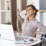 Portrait of young stressed woman sitting at home office desk in front of laptop, touching aching back with pained expression, suffering from backache after working on laptop