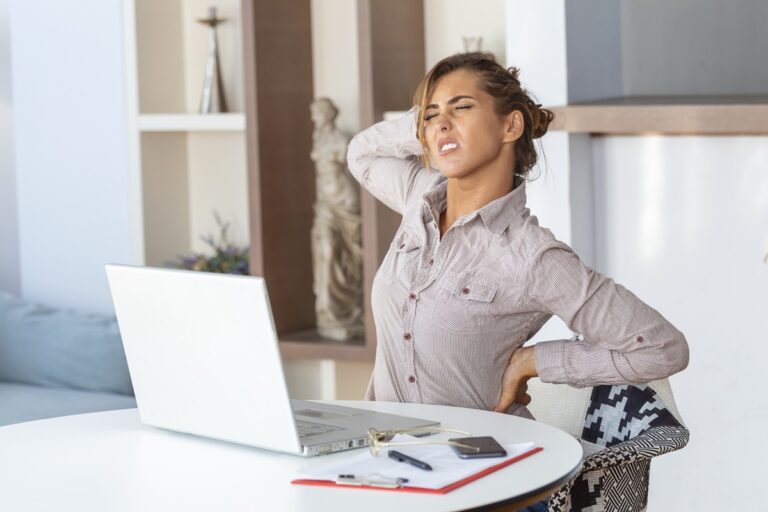 Portrait of young stressed woman sitting at home office desk in front of laptop, touching aching back with pained expression, suffering from backache after working on laptop